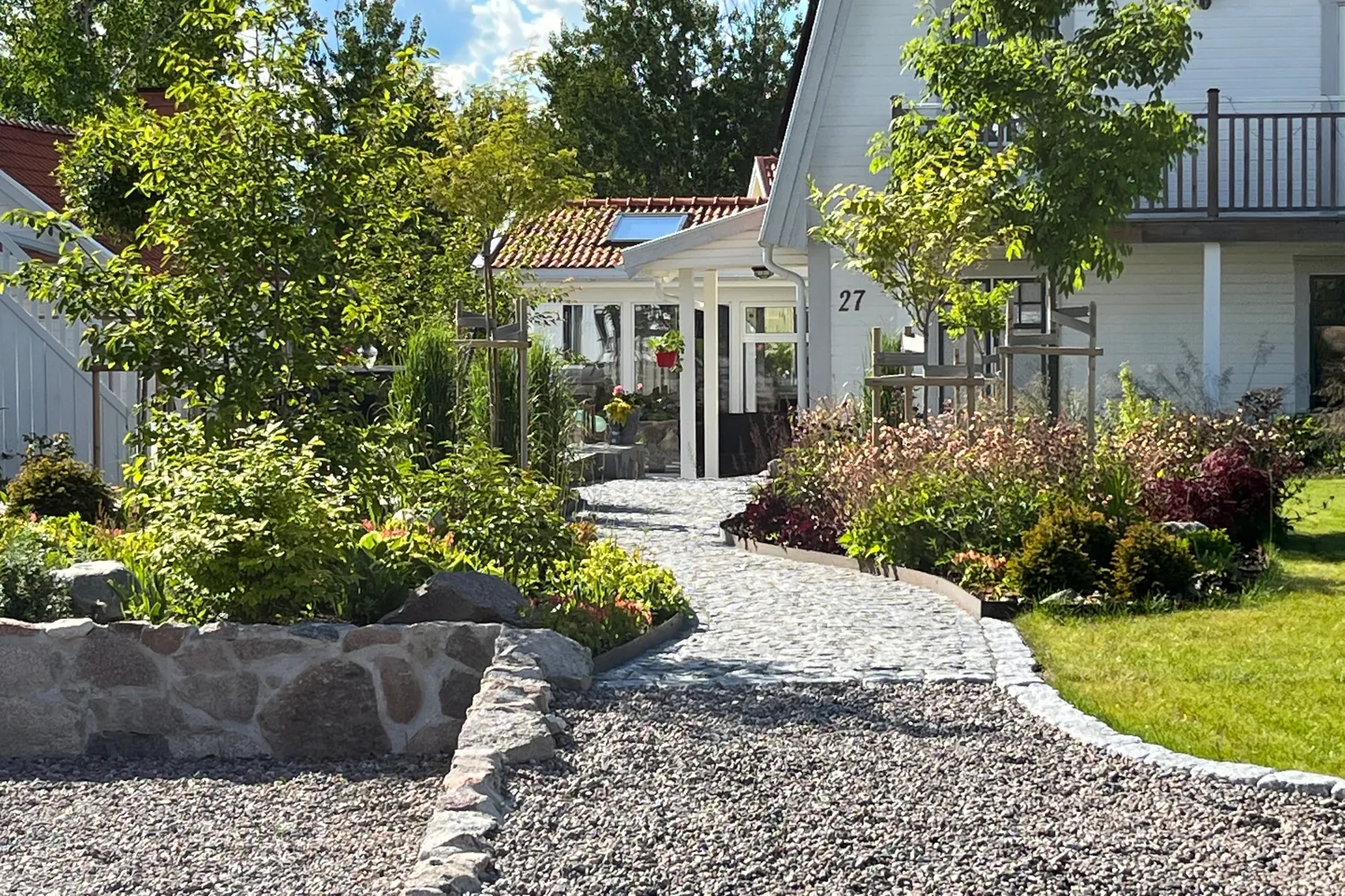 Smågatstensgång svänger mellan nyanlagda rabatter och en mur av natursten fram till en vit villa med inglasad veranda